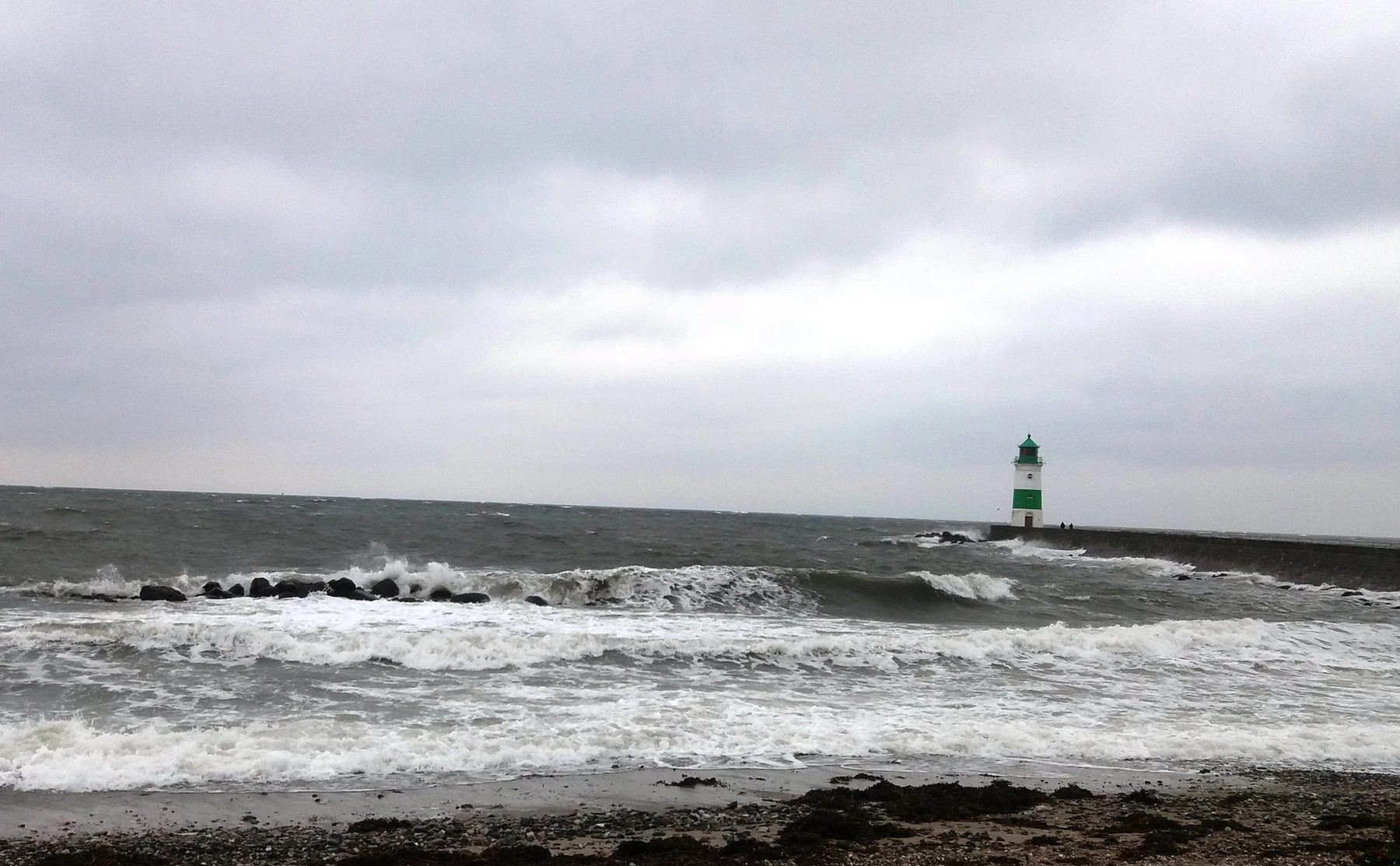 Green-and-white lighthouse on its mole, with rough grey water and waves breaking over rocks.