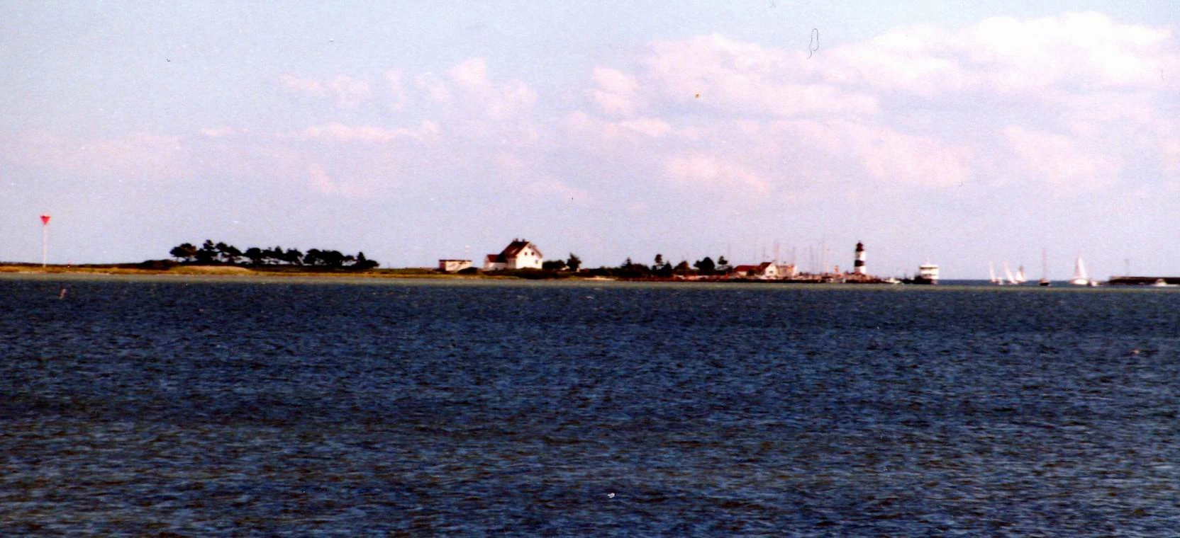 Low coast with the pilots’ house, harbour buildings and a black-and-white lighthouse under a wide sky.